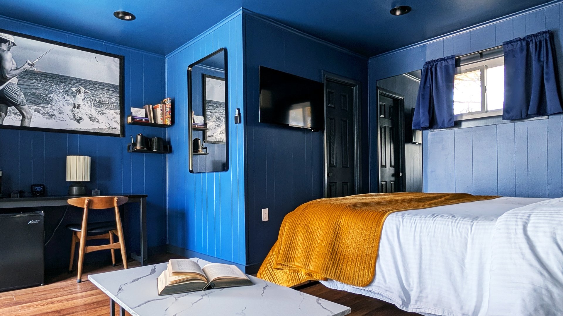 Indigo Studio King opposite wall with solid deep blue paneling, black-and-white surfer photograph, mustard throw, and desk nook